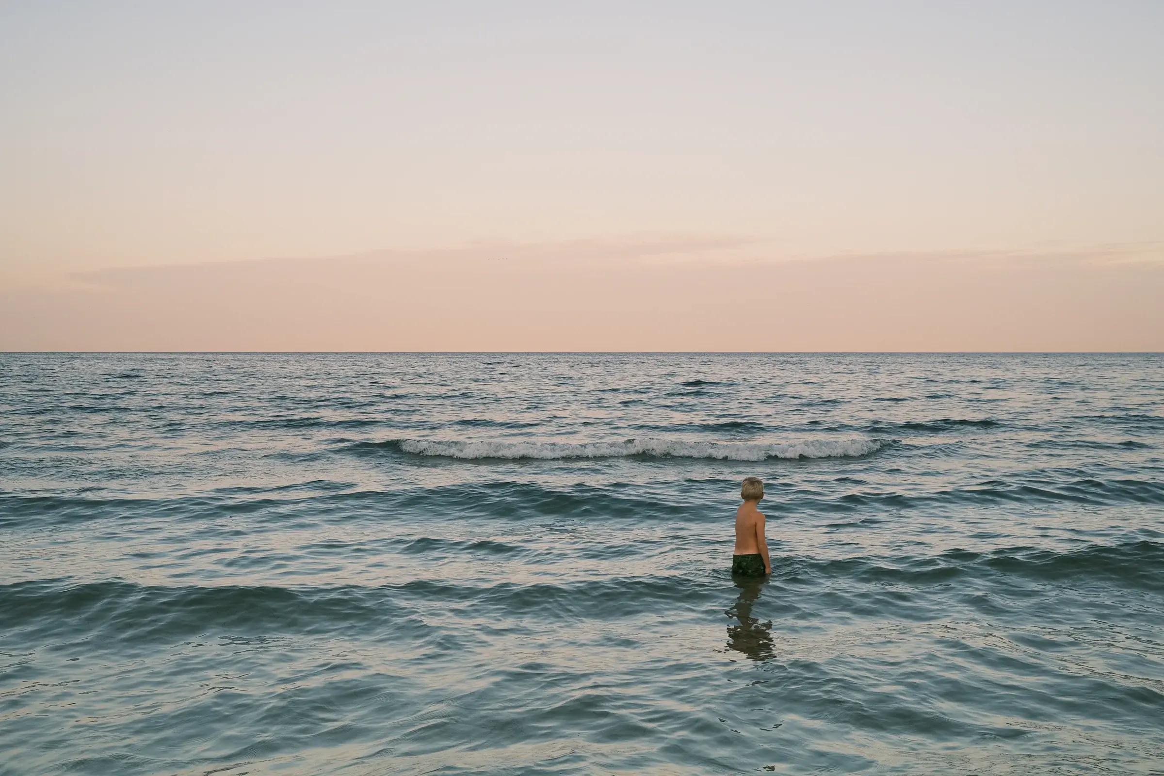 Person standing waist-deep in ocean waves at sunset, with calm horizon and pastel sky.